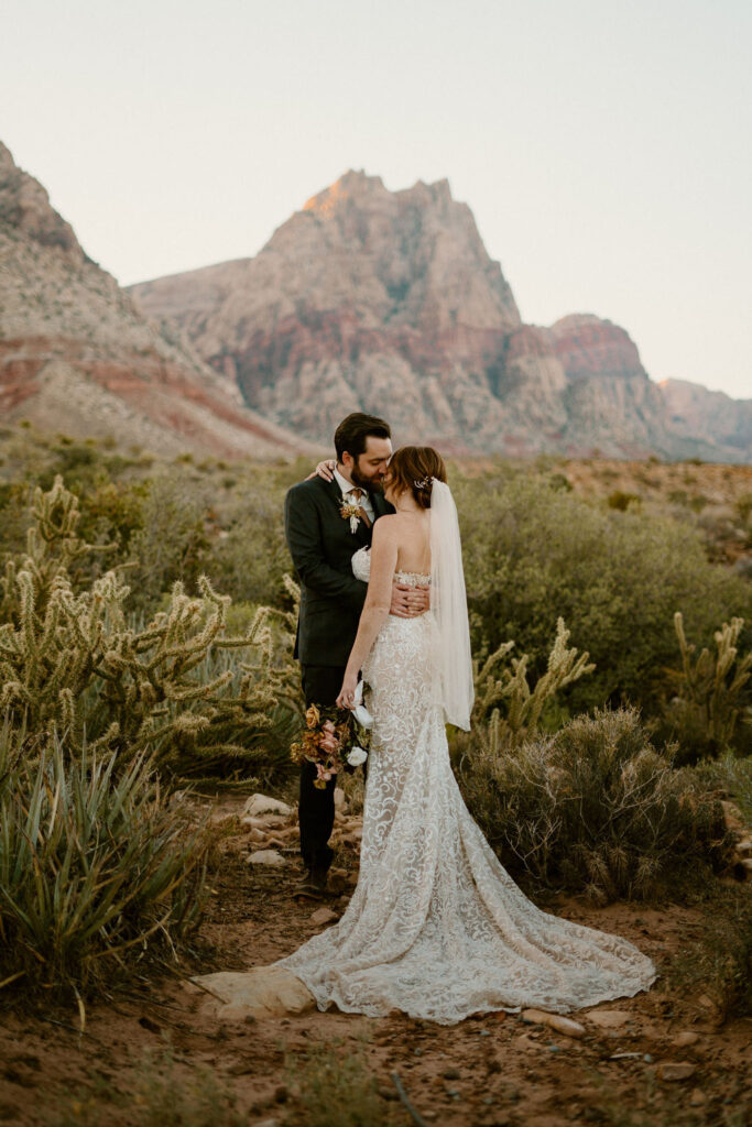 Groom and bride going in for a kiss. Surrounded by dessert greenery and the brides train cascading on the desert ground. 