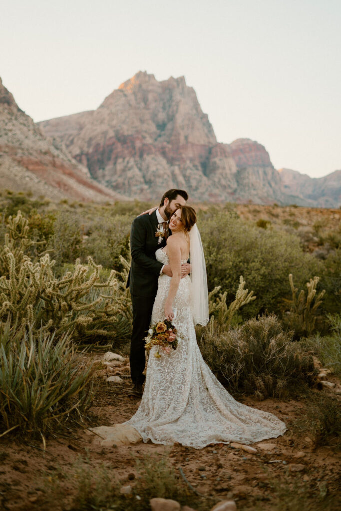 Groom kisses his brides cheek as she smiles and her beautiful gown cascades on the desert ground. Red Rock Canyon mountains in the background. 