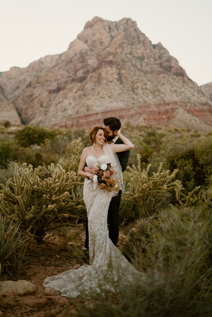 Bride pulls grooms neck into her as he holds her waist. Surrounded by beautiful desert greenery and mountain in background. 