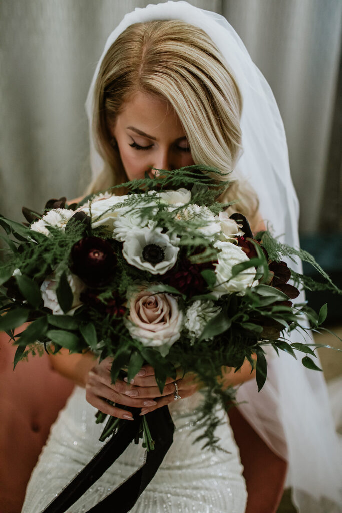 Bride holding a dark and moody styled bouquet. It is at he face and she looks down. A veil pinned into her hair. 