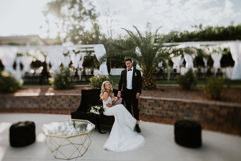 Blurry photo of bride and grooms reception in background. Bride and groom central focus. Groom is standing holding brides hand as she sits on a dark black velvet chair. Her bouquet behind her and dress cascading on the ground. 