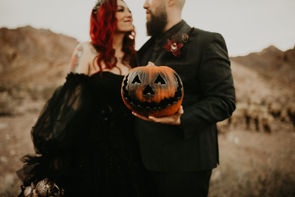 Bride and groom wearing all black. The groom is holding a smiling carved pumpkin. The cut out eyes and smile is blacked. 