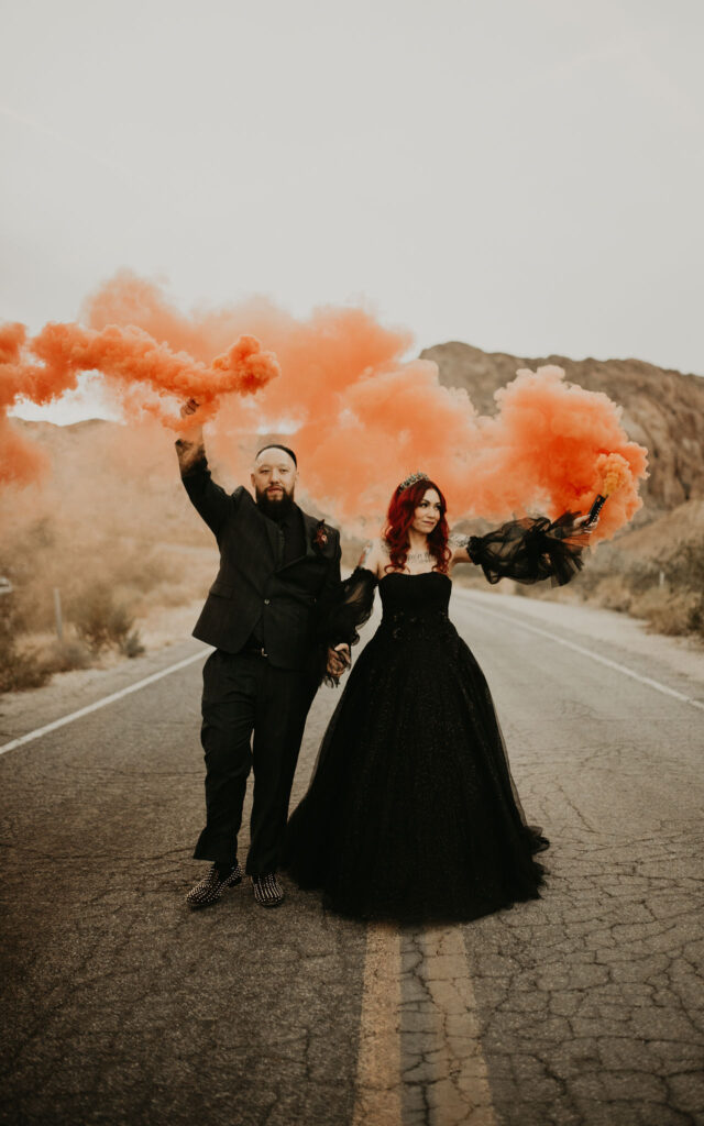 Bride and groom in all black outfits. Holding bright orange smoke grenade walking along the road. 