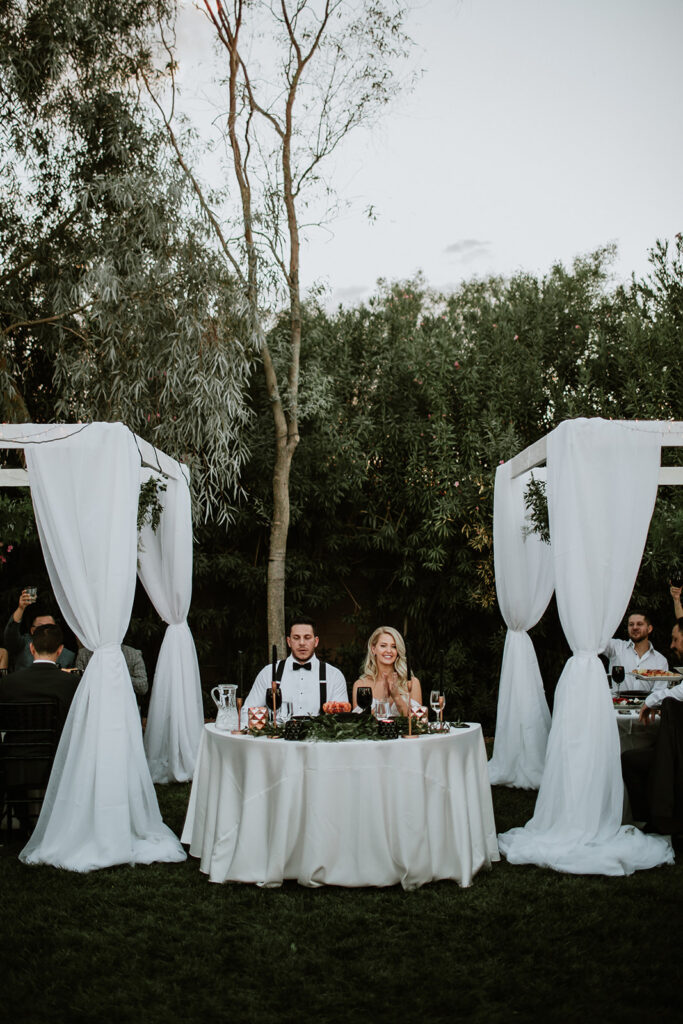 Bride and groom sit at a round Sweetheart table. The smile with lush greenery behind them. Dark and moody table decor decorates their table. Guest cheer to the right and left. 