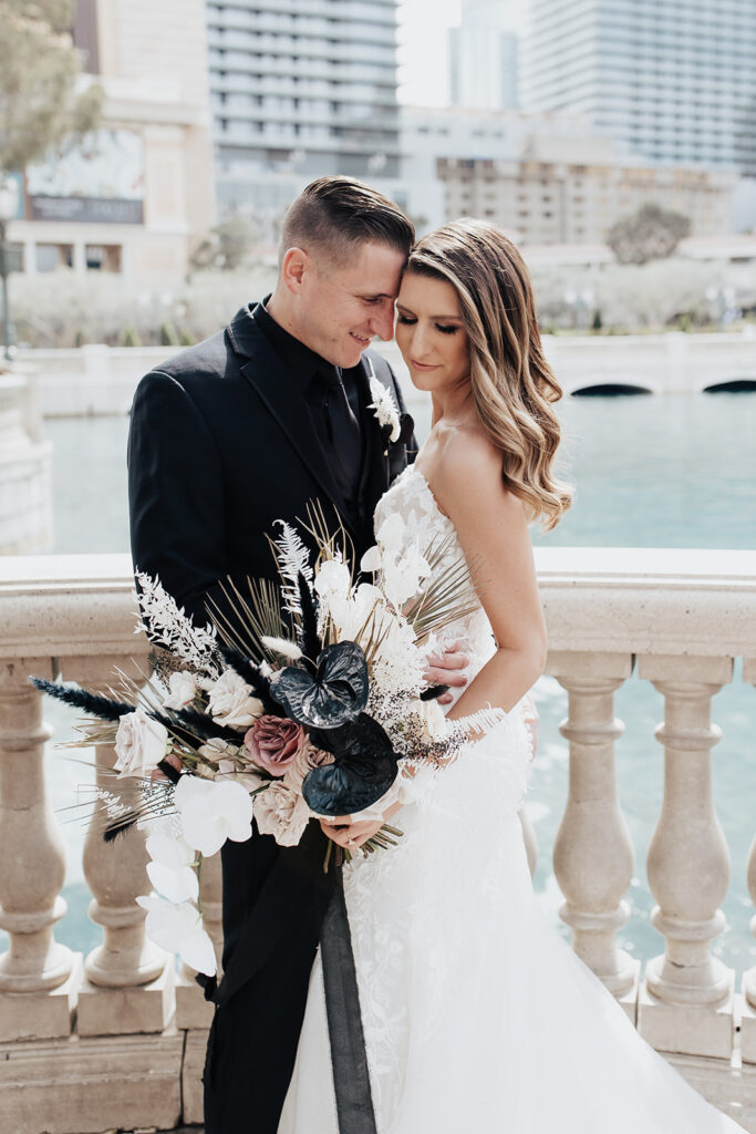 Bride and groom standing in front of the Bellagio fountains. Their heads pressed together. The grooms striking black suit contrasts perfectly with the brides dark and moody bouquet. 