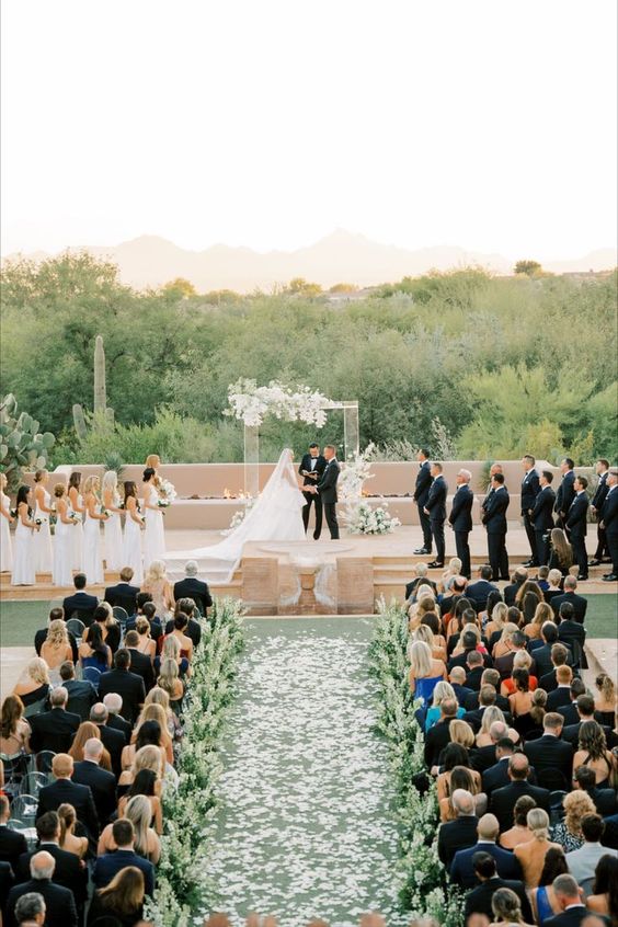 Aisle: Circle Or Runway? Beautiful runway aisle with florals and greenery. Filled with guests. Bride and groom at the center. 
