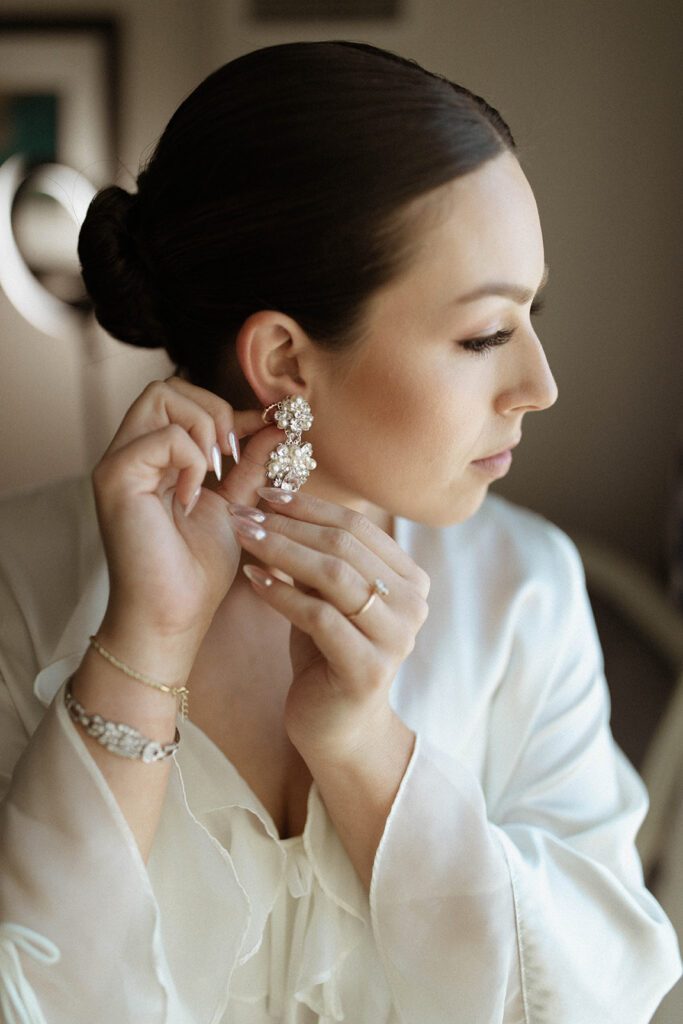 Bride has a slicked back elegant bun styled in her hair. She is putting on her diamond dangling earing while wearing her silk bride robe to finish getting ready. 