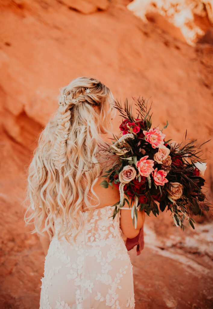 Bright blonde haired bride shows us her intricate braided and curled styled hair. She holds a big maroon and red wild flower bouquet. 