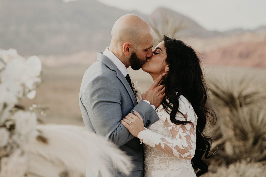 Just The Two Of Us Elopement in the Desert Bride and Groom share a kiss. Her long brunette hair curls all the way down her back. Groom caresses her hair. She wears a long floral lace sleeves.  Pampas is blurry in the forefront of the photo.