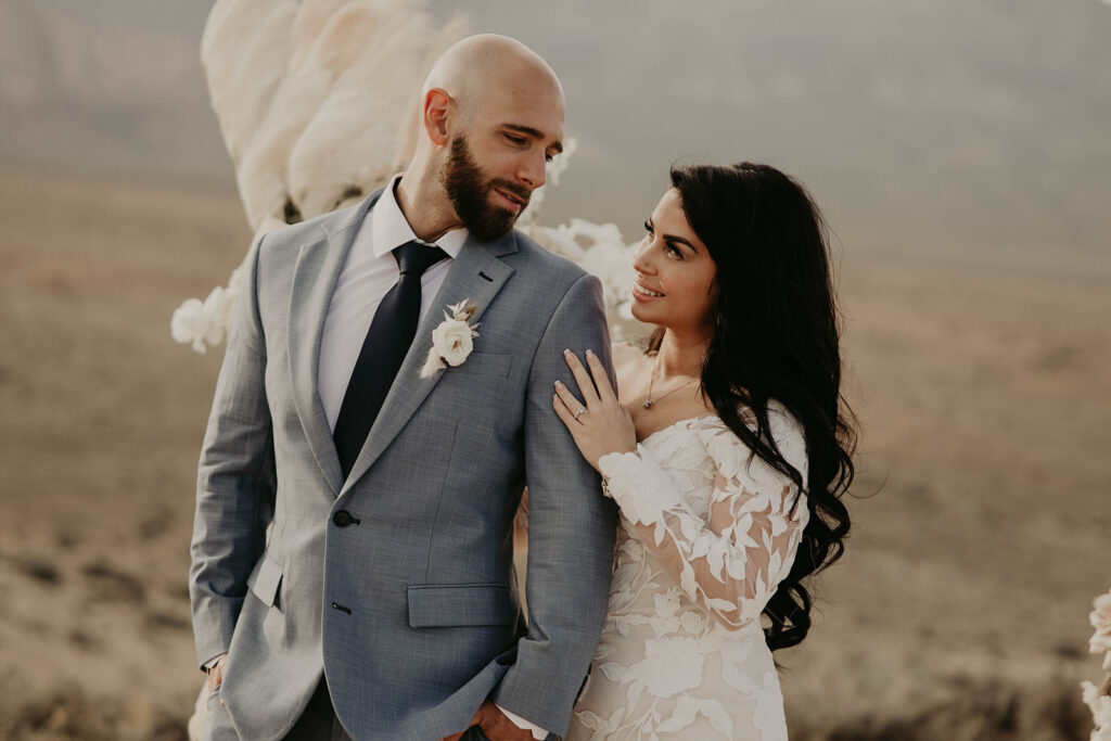 Just The Two Of Us Elopement in the Desert groom in blue grey suit smirks at his bride as she caresses his arm with a wide smile. Her brunette hair curls down her back with a long sleeve floral lace wedding dress. 