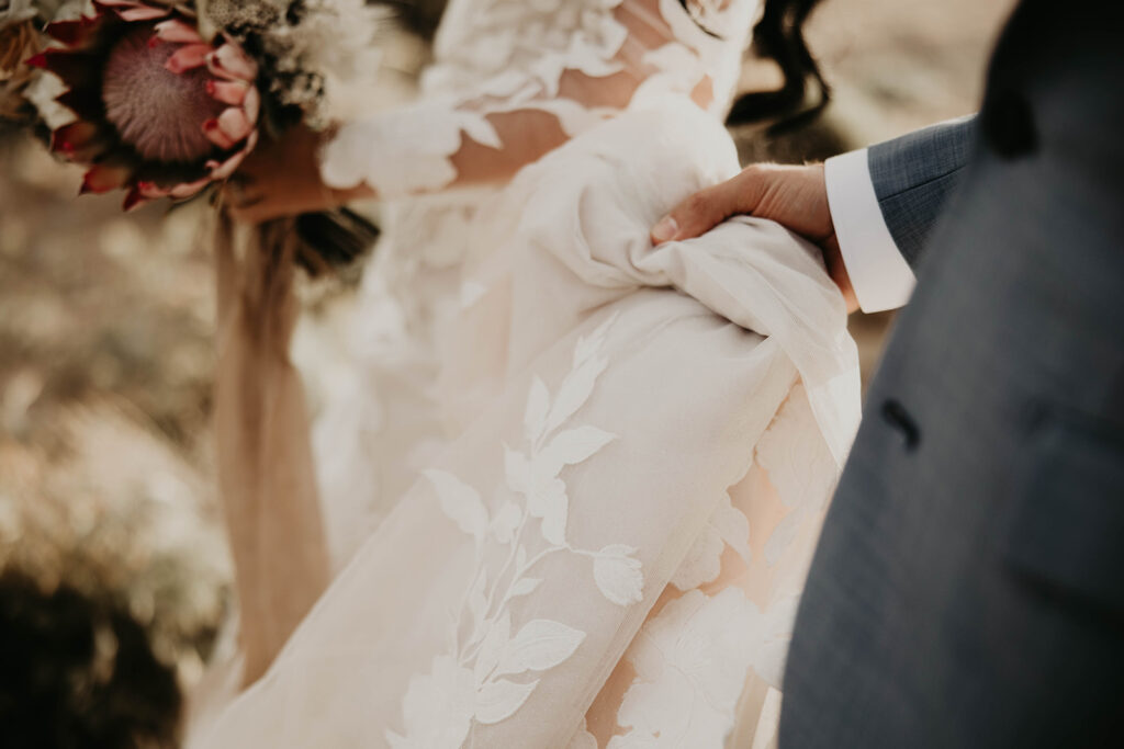 Groom is holding his brides train as the camera focuses on the floral lace decorated on her dress. 