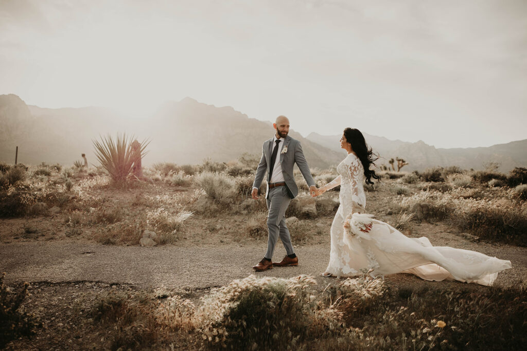 Just The Two Of Us Elopement in the Desert. Bride and groom walk along Red Rock Canyon Trail. Groom is wearing a tailor made blue grey suit. The bride has an elegant mermaid style dress with floral lace sleeves. Her train flowing in the wind behind her. 