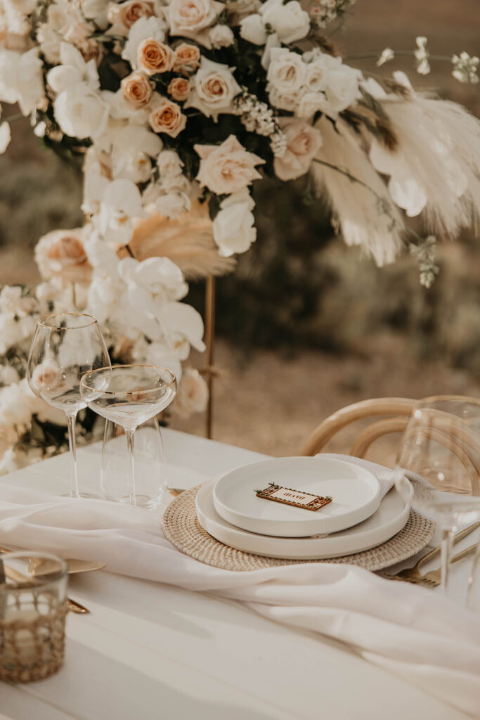 Close up of the sweetheart table. White plates with boho styled ground arch in the background. The ground arch has pampas, roses of white, cream, toffee, and peach decorated throughout. 