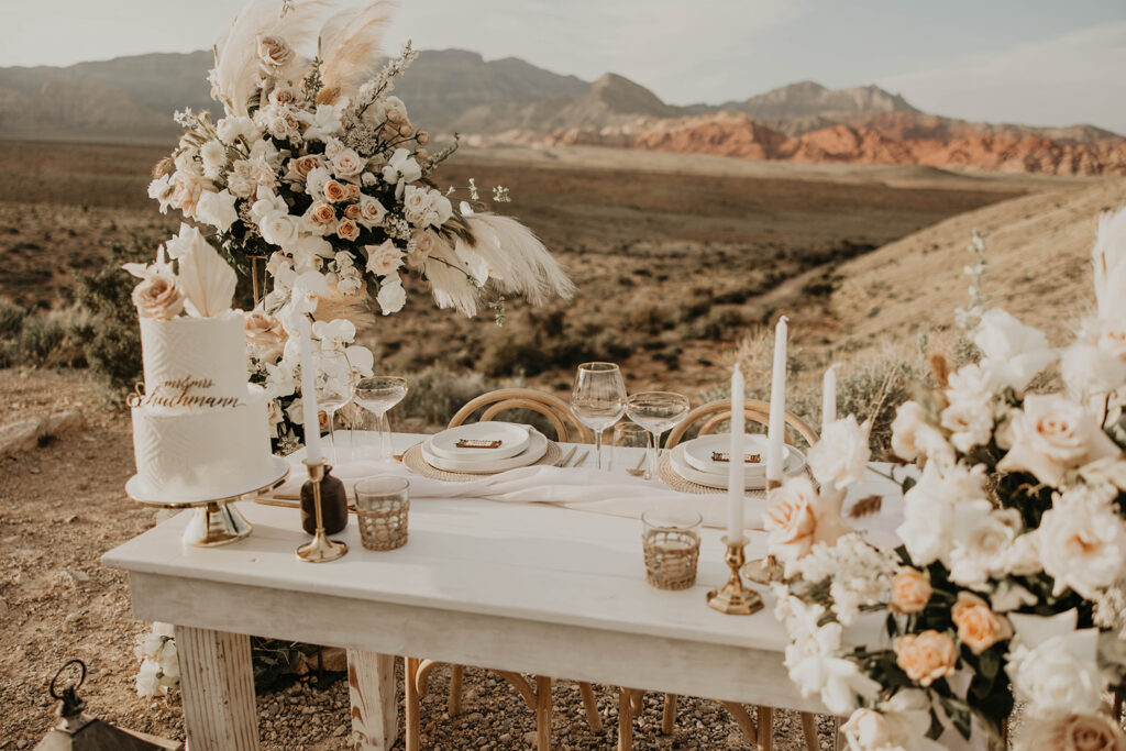 Decorated sweetheart table in the Red Rock canyon. White, cream, coral, and toffee colored boho floral arches decorate the white wooden table. A two tiered white wedding cake sits on the corner of the table. White plates and candles are placed for the couple. 