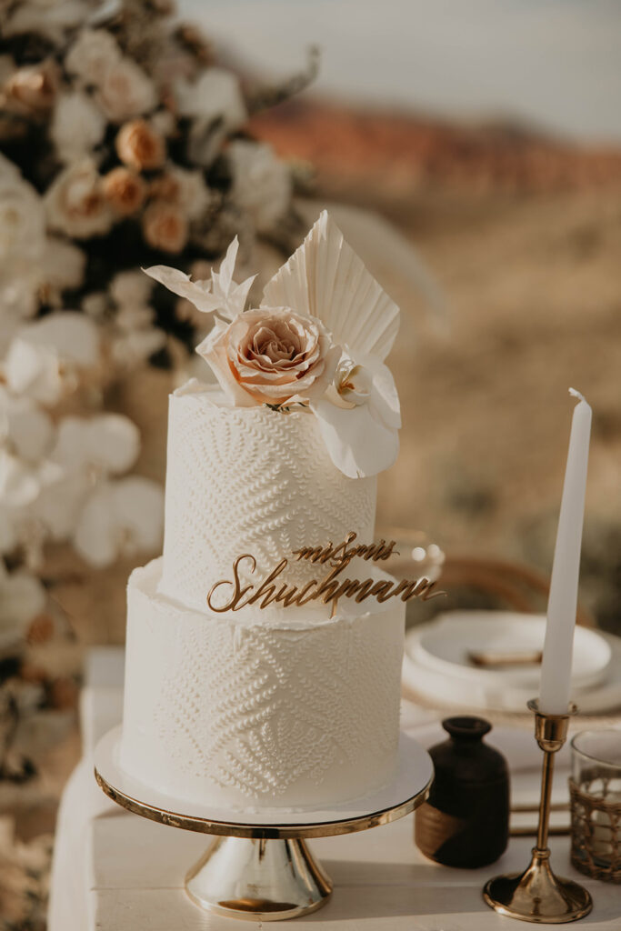 Close up of two tiered wedding cake with intricate leaf design. A boho stlye floral decoration lays on top of the cake with a golden personalized cake topper.