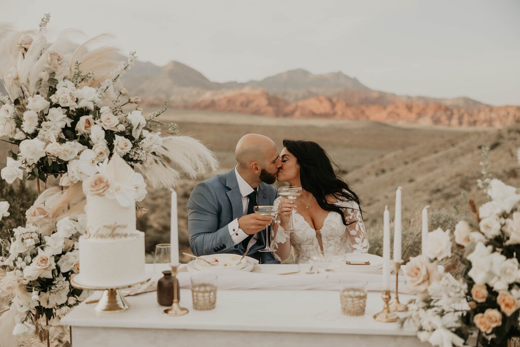 Just The Two Of Us Elopement in the Desert Bride and groom sit at the sweetheart table. Sharing a kiss as they clink their champagne glasses. The grooms plate holds little crumbs from the  two tiered white wedding cake. 