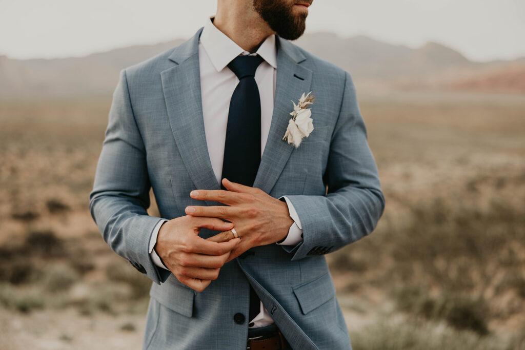 Just The Two Of Us Elopement in the Desert. Shot of grooms torso. He twirls his wedding ring looking off into the distance. His blue grey suit tailored perfectly with a deep navy blue tie. 
