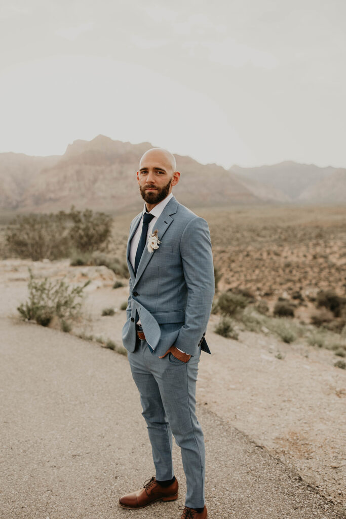 Wide shot of just the groom. Red Rock canyon mountains in the background make the blue grey tailored suit pop against the red and green rock. The groom has his hands in his pocket with a perfectly trimmed beard. 
