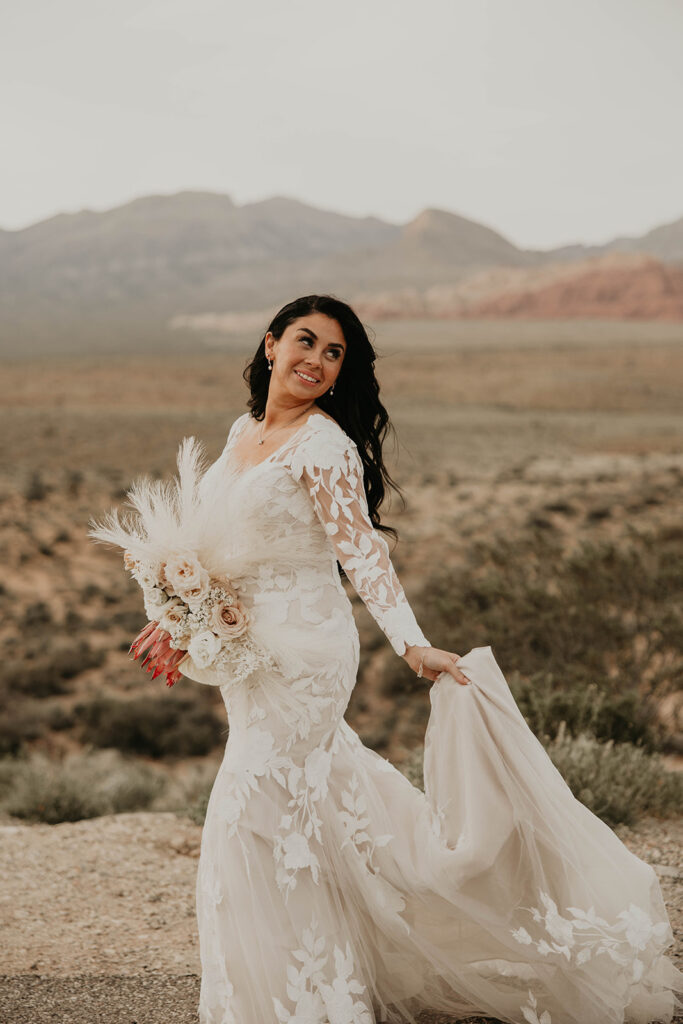 Just The Two Of Us Elopement in the Desert. Brunette bride holding her mermaid dress train. Her long hair curled down her back and her beautiful boho styled bouquet with bright pink Protea. 