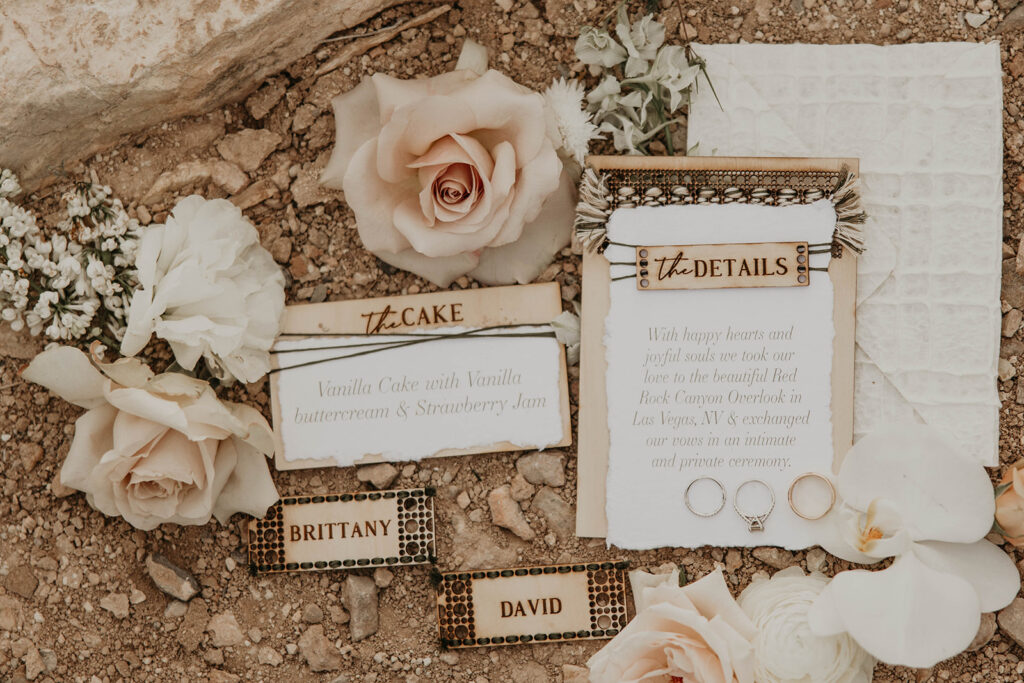 Just The Two Of Us Elopement in the Desert. Close up of the couples stationary and jewelry. The stationary has been detailed out on wooden pieces with soft pink roses surrounding.