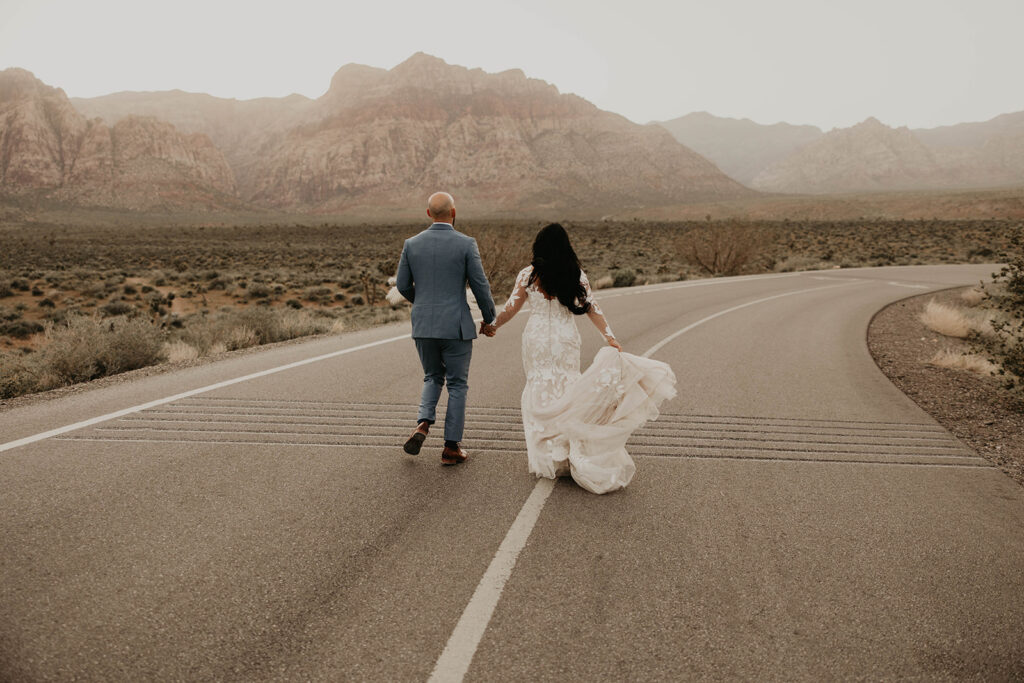 Groom in tailormade blue grey suit and bride in a mermaid floral lace wedding dress run along a back road in Red Rock Canyon. 