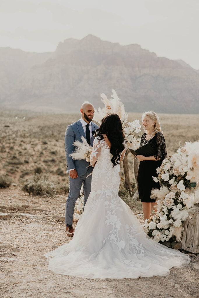 Bride and groom standing at Red Rock Canyon overlook. The brides train flows out behind her. Boho white and cream ground arches stand to the side as the officiant is marrying the couple. 