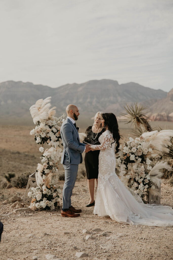 Officiant standing in the middle of boho styled ground floral arch. Bride and groom stare at each other holding hands. Groom wears a tailor made blue grey suit. Bride is wearing a floral lace mermaid dress. 