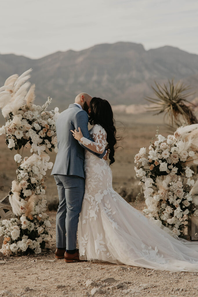 Groom in blue grey suit and bride in a floral lace mermaid dress with long train kiss in the middle of their boho styled floral ground arch. 