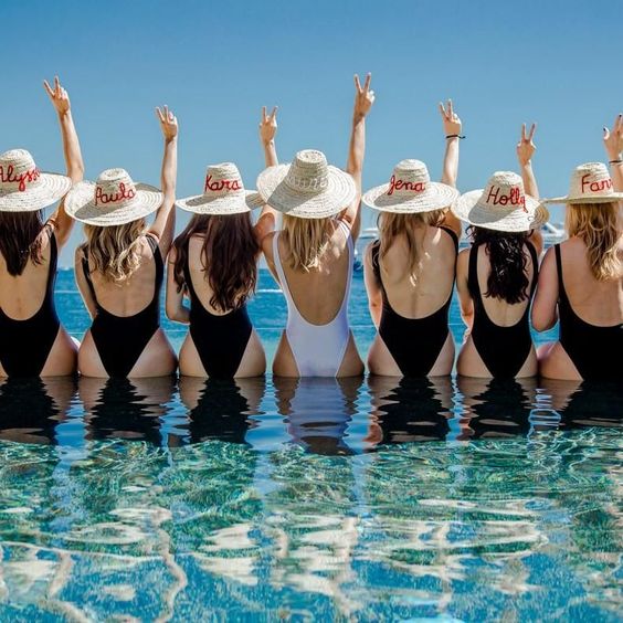 5 Trendiest Bach Party Themes Bride and bridesmaids stand in a pool overlooking the ocean. The party wears matching one piece swimsuits. The bride wears white while everyone else is in black. They have personalized sun hats and have a peace sign up in the air. 