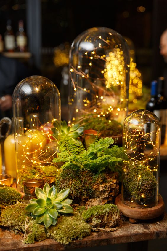 Themed table setting of moss and greenery surrounding glass tubes with twinkle lights inside. 