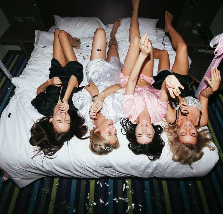 5 Trendiest Bach Party Themes Bride and bridesmaids laying on a bed, confetti all around them. The bride wears a white silk pj while the bridesmaids wear pink and black matching pjs. 