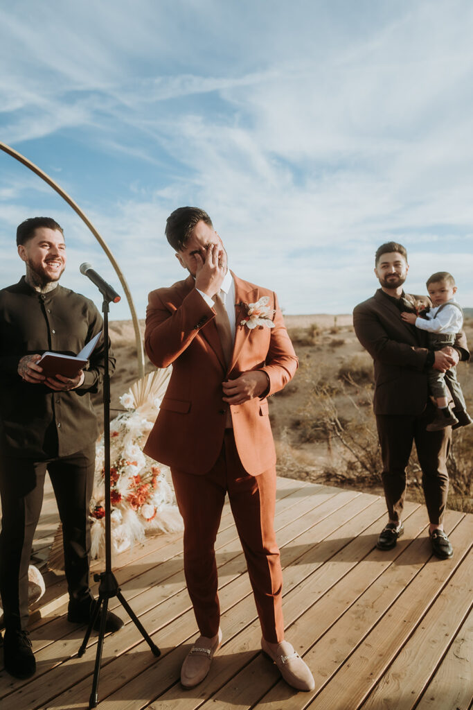 The groom in a burnt orange suit with suede shoes holds his eyes as he is overcome with emotion looking at his bride for the first time.