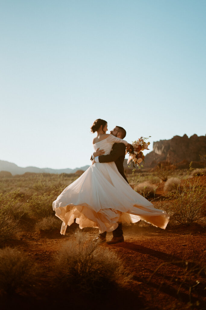 Fun & Creative Wedding Day Photo Poses and Ideas brides wears an off the should long flowy wedding dress. The groom holds her in his arms and spins her around letting her dress twirl perfectly. 