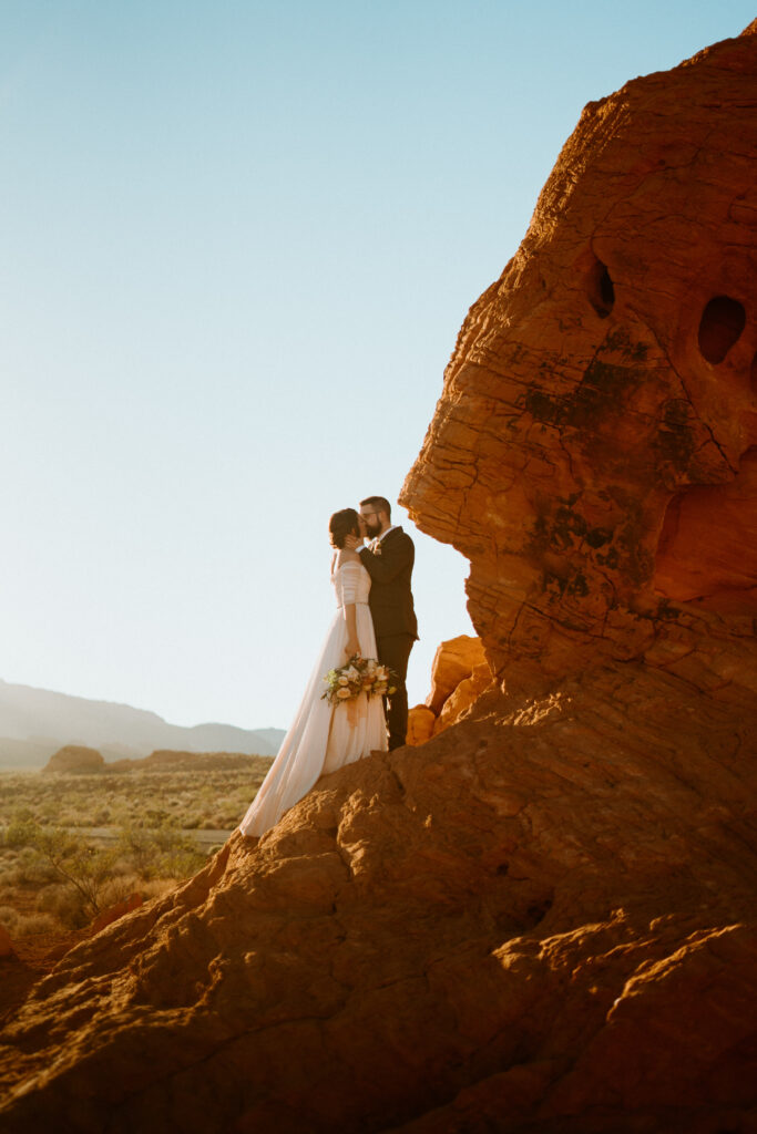 Fun & Creative Wedding Day Photo Poses and Ideas Bride and groom stand on top of a red rock, the sun setting behind them sharing a kiss in a dramatic wide angle shot. 