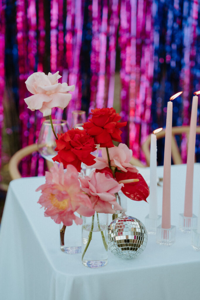 Pink and red roses is small glass vases. With a tiny disco ball next to pink and white candles. White table clothes with pink, red, and purple center pieces. 