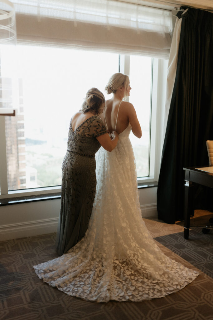 Mother of the bride helps her button of her lace wedding dress with open back. 