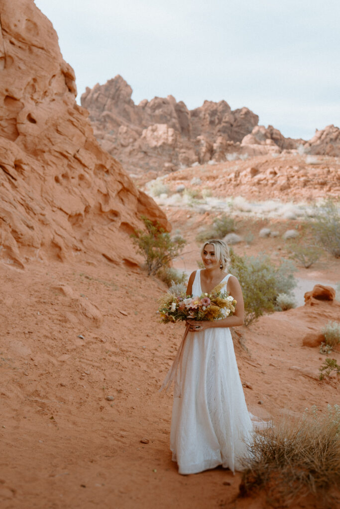 Fun & Creative Wedding Day Photo Poses and Ideas the bride in an elegant halter white wedding dress. Walks up the red rock pathway holding a pink, peach, and white bridal bouquet. 