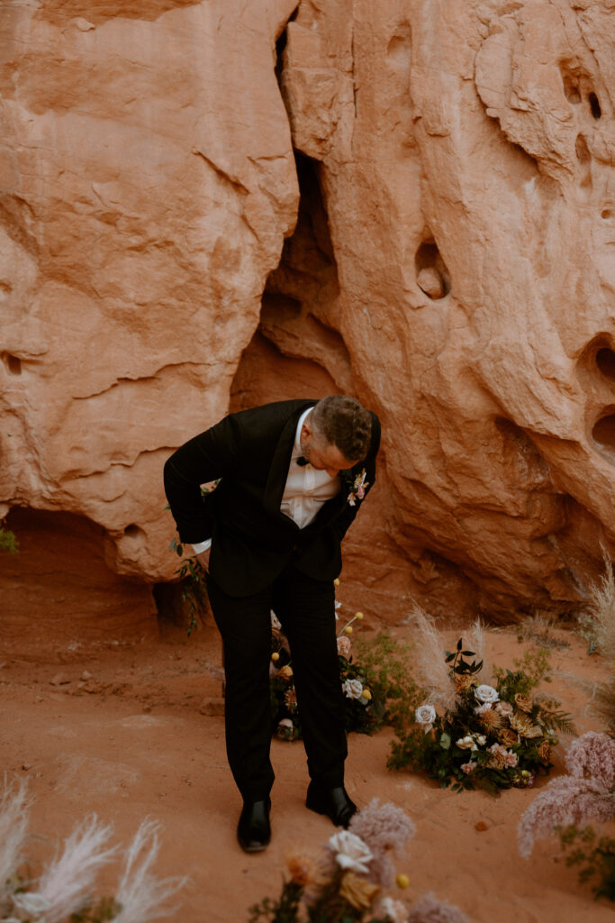 The groom, in his black tux, looks down overwhelmed with emotion watching his bride walk towards him. Ground floral arches surround him. 