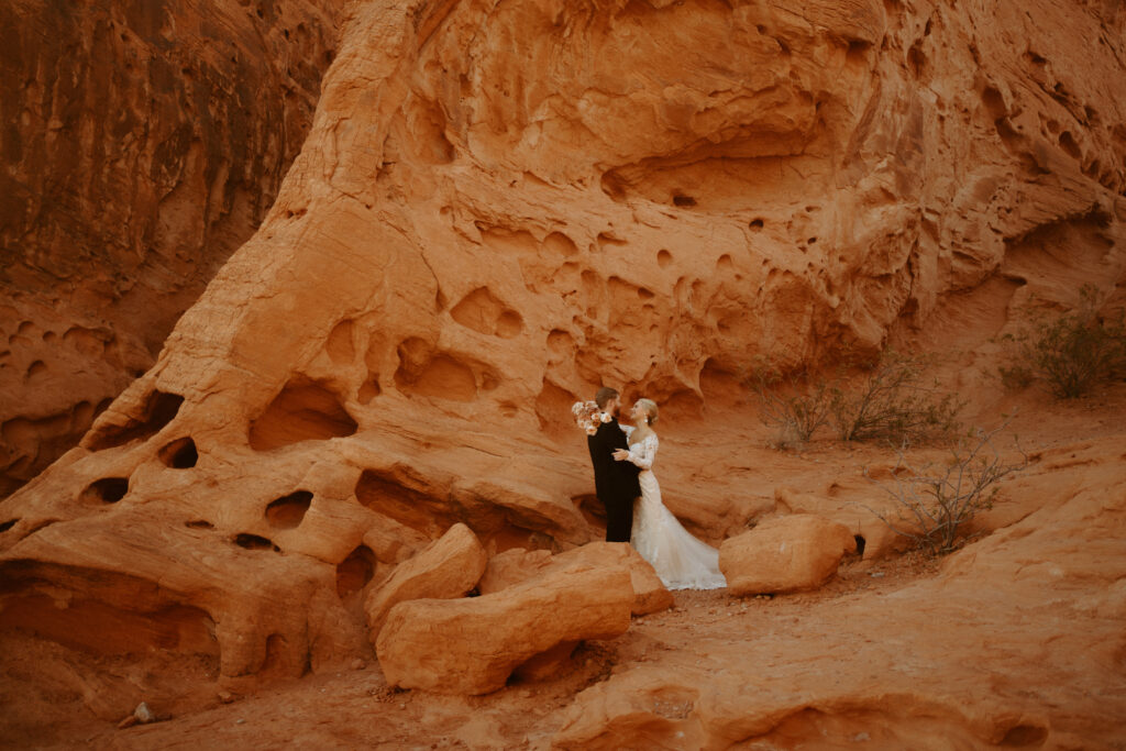 Las Vegas has the Craziest Winds! Wide dramatic shot of couple against the red rock. Groom is in an all black suit, bride is wearing an off the shoulders fitted lace wedding dress. With long sleeves and long lace train. 