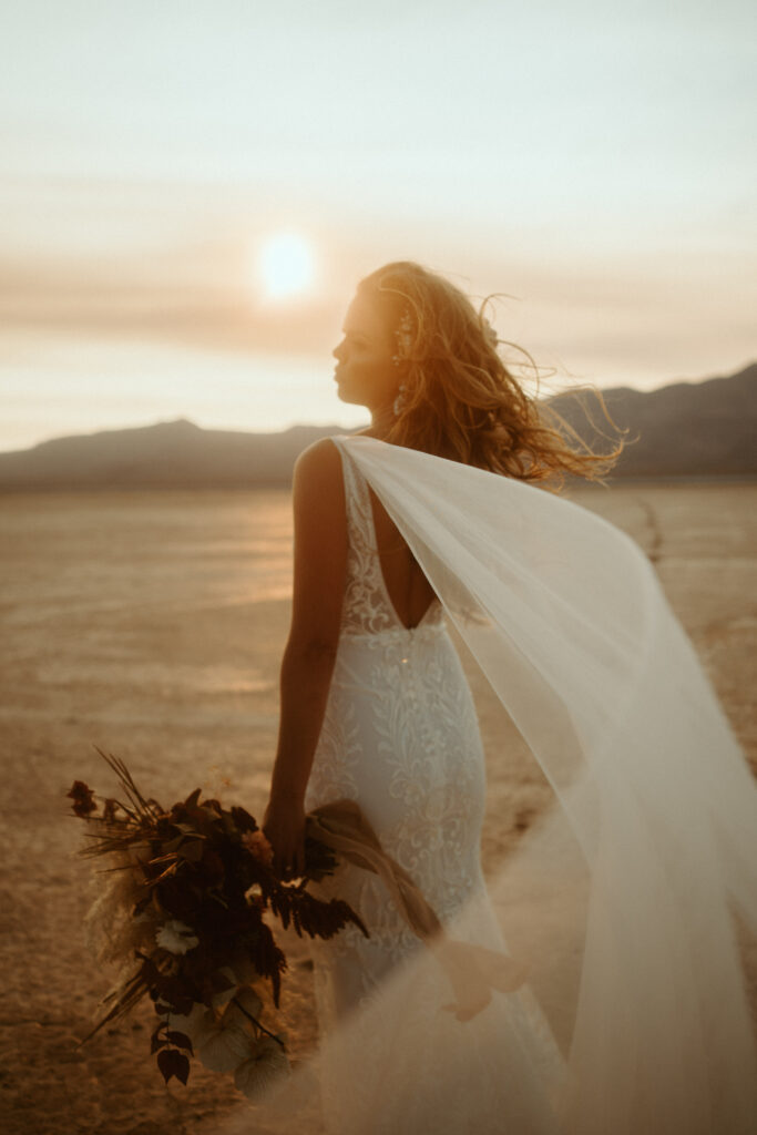 Fun & Creative Wedding Day Photo Poses and Ideas The brides shoulder train blows in the wind and the sun is directly behind her giving her a soft silhouette image. She holds her large boho styled bouquet down at her side. 