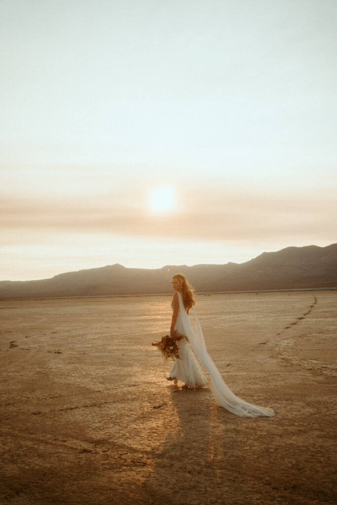 The Ultimate Guide to Different Types of Wedding Dresses: From Classic to Contemporary. Wide angle shot of bride walking in the Las Vegas desert. The sun is blocked by clouds. The bride has a fitted lace wedding gown with a long flowing shoulder veil. 