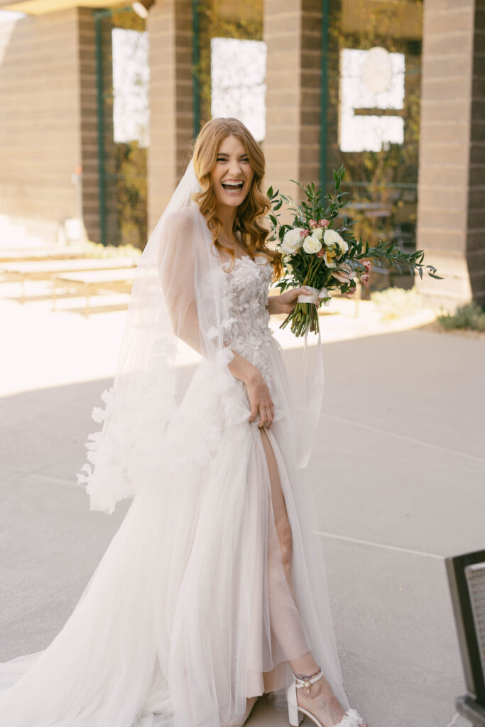 The Ultimate Guide to Different Types of Wedding Dresses: From Classic to Contemporary. Bride laughing as she shows off her contemporary wedding dress with high leg slight and fitted bodice. 