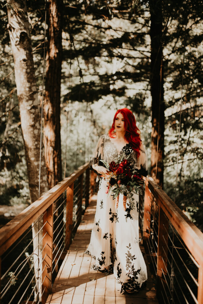 Fun & Creative Wedding Day Photo Poses and Ideas bride in a unique black and white wedding dress, with her striking red hair and bright red and green bridal bouquet to match, stands on a boarded walkway in the middle of a forest. 