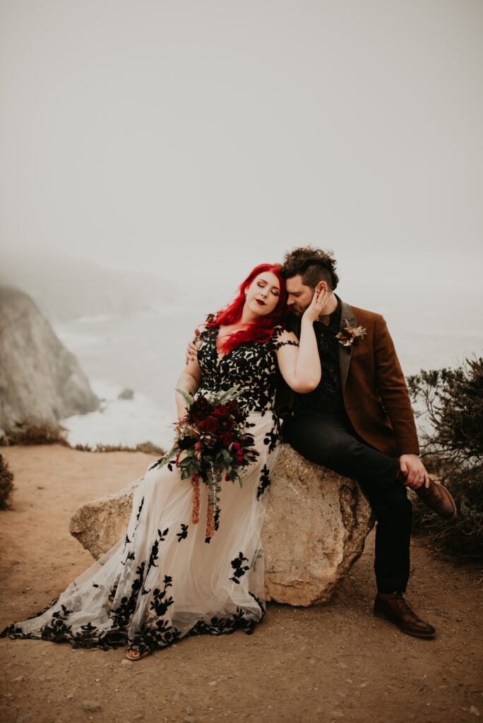 Bride in her white and black wedding dress, holds her grooms face softly. They sit on a rock at the edge of a cliff. Directly behind them is the ocean. 