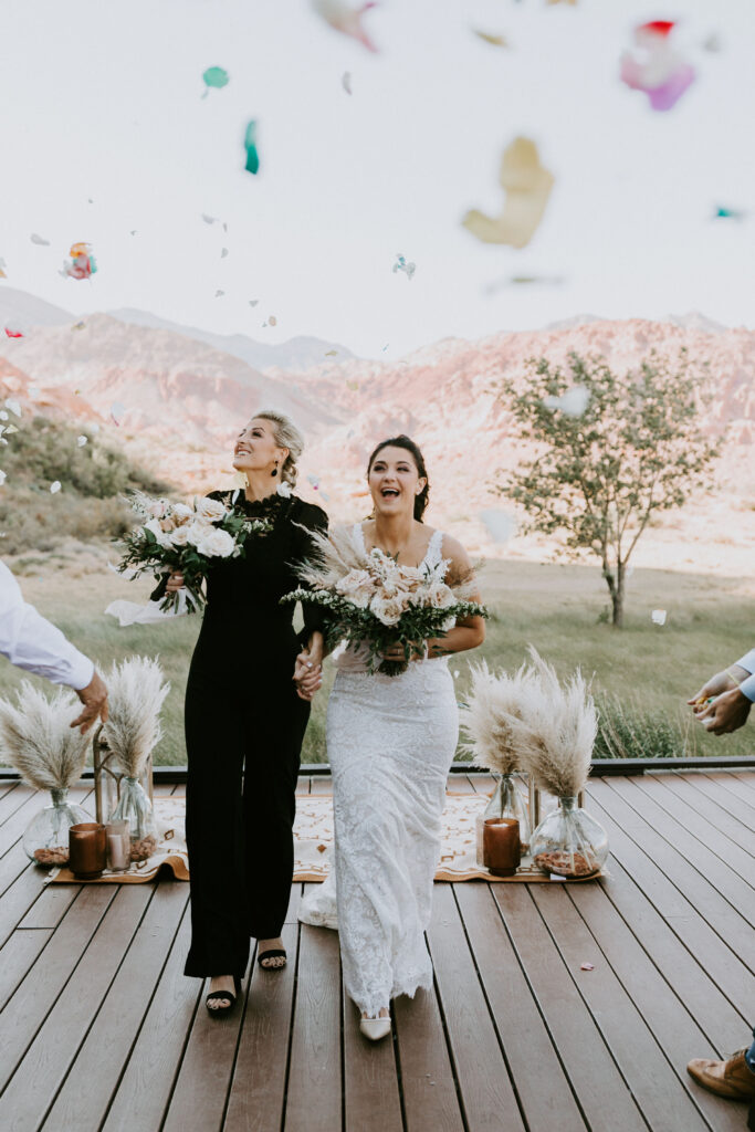 Bride in a black jumpsuit and bride in a halter fitted lace dress walk hand in hand as flower petals rain all around them. 