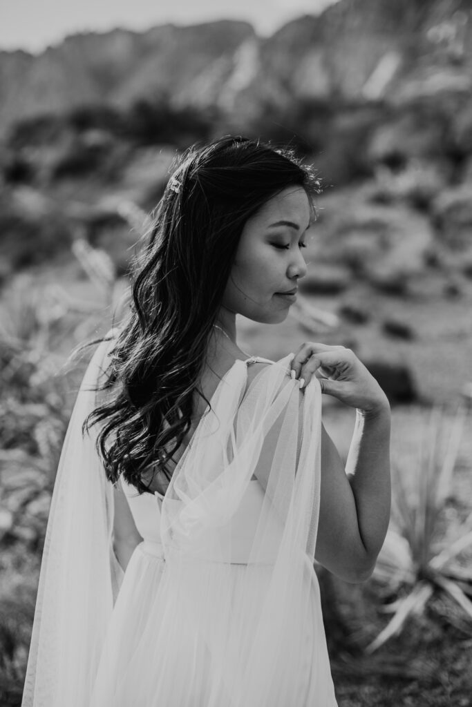 Love Unleashed: Colorful Las Vegas Elopement. Black and white photo of the bride turned away from the camera holding her shoulder cape. The bride wears a V-neck silk top with a long shoulder cape and tulle flowy skirt