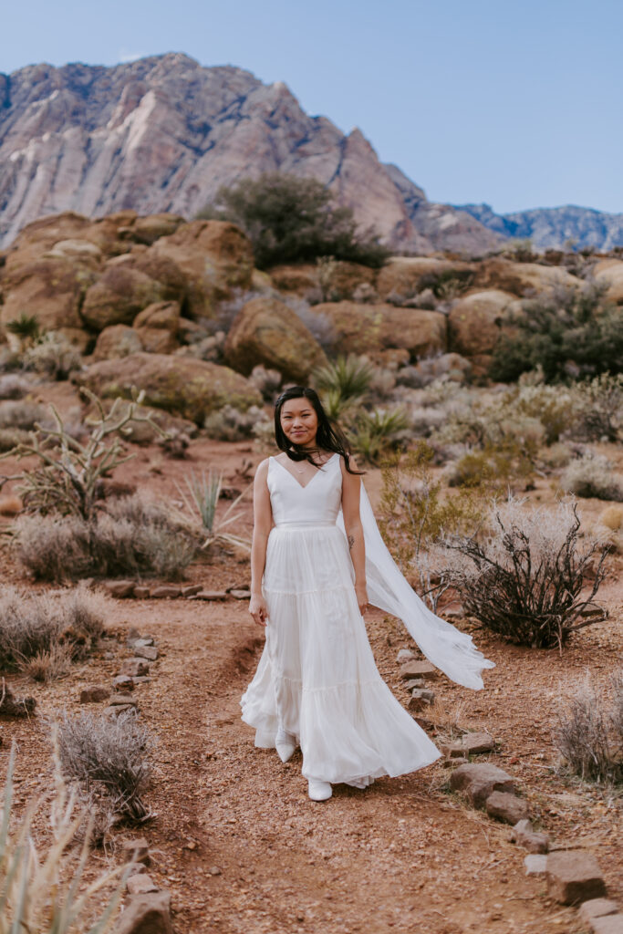 Love Unleashed: Colorful Las Vegas Elopement  The bride wears a V-neck silk top with a long shoulder cape and tulle flowy skirt. She walks down a desert path with the wind blowing her shoulder cape. 