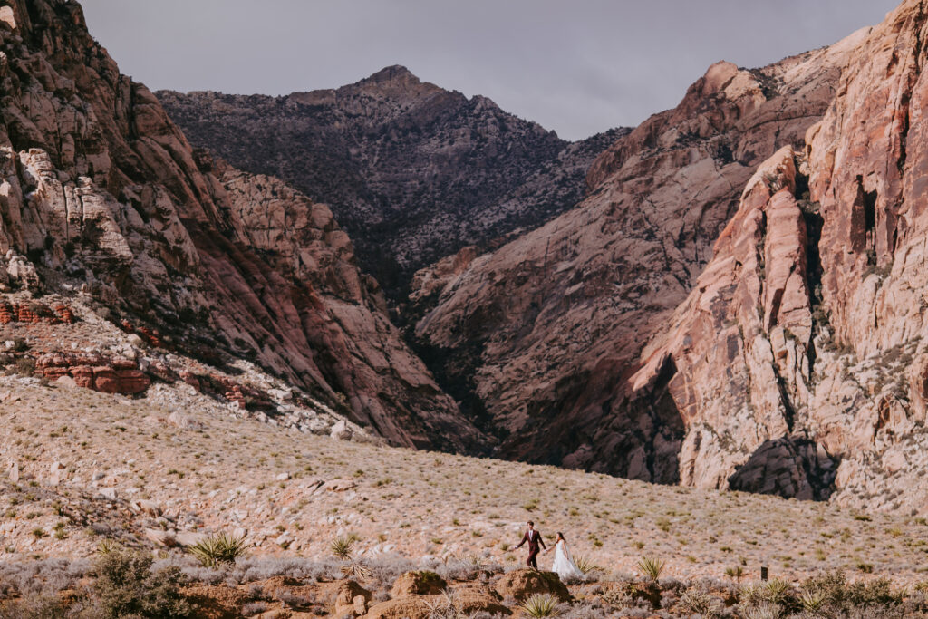 Love Unleashed: Colorful Las Vegas Elopement. Wide angle dramatic shot of the Las Vegas Red Rock mountains with the bride and groom visible in the far distance. 