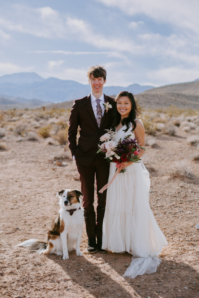Love Unleashed: Colorful Las Vegas Elopement  The bride and groom take a family photo with their dog as he sits perfectly looking at the camera. The bride wears a V-neck silk top with a long shoulder cape and tulle flowy skirt/ The groom wears a deep maroon suit with purple paisley tie. 