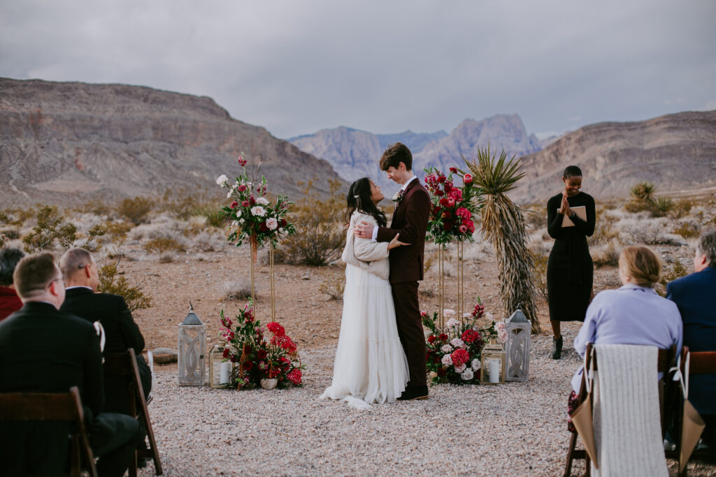 Love Unleashed: Colorful Las Vegas Elopement. The couple stands at their maroon, purple, and pink floral arch with the desert mountains in the background. The bride wears a V-neck silk top with a long shoulder cape and tulle flowy skirt. The groom wears a deep maroon suit with purple paisley tie. 